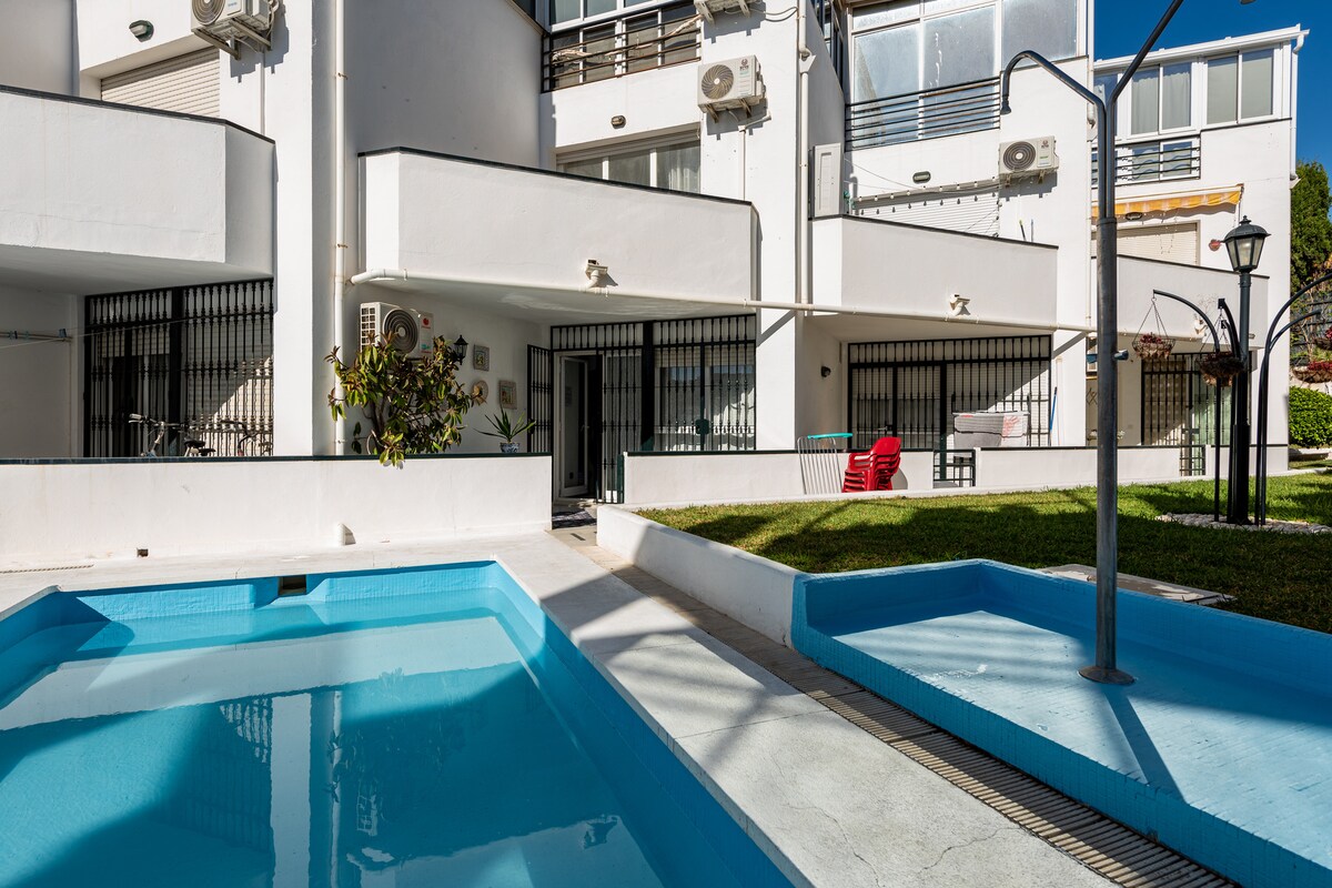 An inviting outdoor area features a two-tiered pool surrounded by well-maintained greenery. The light-colored building showcases large glass doors, offering views into the interior spaces. Red lounge chairs are positioned for relaxation near the pool, enhancing the communal atmosphere.