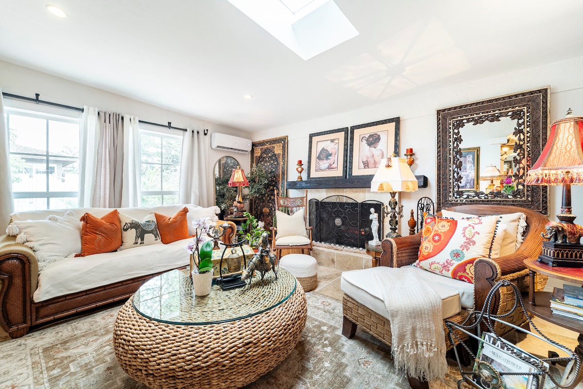 The living room features a comfortable seating area with a woven coffee table at its center. Natural light filters through large windows, enhancing the inviting aesthetic. Decorative art pieces line the walls, and a soft rug adds warmth to the space. A skylight above provides additional brightness.
