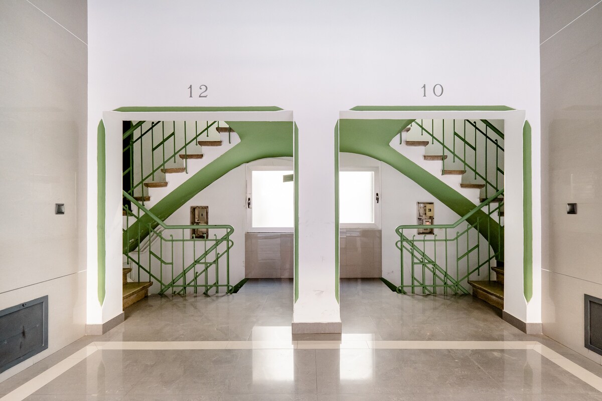A well-lit stairwell features two adjacent staircases, numbered 10 and 12, with green railings and walls. Natural light is provided by windows at the top of each staircase, illuminating the marble floor and creating a clean, inviting appearance.