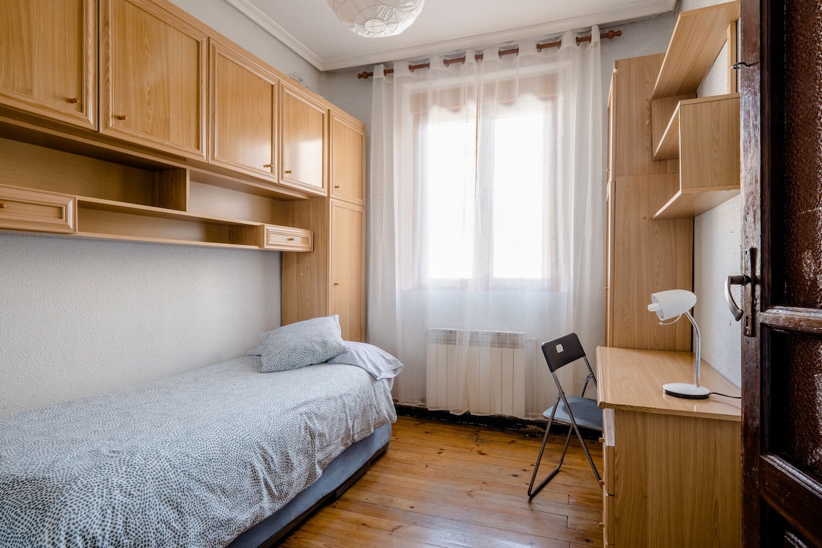 A light-filled private room is presented, featuring a single bed with a soft gray blanket and a pillow. A simple wooden desk and chair are positioned against one wall. Above the bed, wooden shelving offers storage. Natural light filters through large, sheer curtains covering the window.