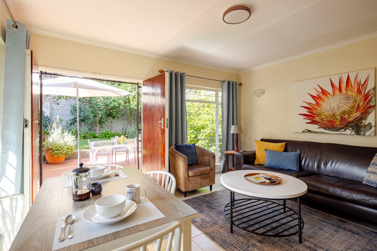 A cozy living space features a comfortable sofa facing a vibrant wall art piece. A dining table set for two is positioned nearby, with a French press and dishes arranged neatly. Large glass doors open to a private garden patio filled with greenery.