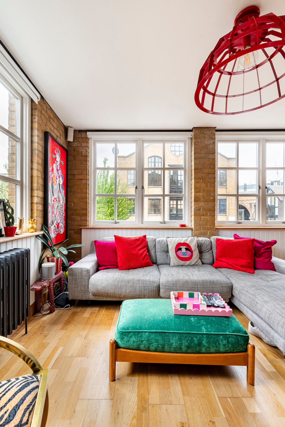 A stylish living area features a grey sectional sofa adorned with vibrant red pillows. A green ottoman rests in front, accompanied by a coffee table displaying colorful magazines. Natural light flows through large windows framed by exposed brick walls, creating a bright atmosphere.
