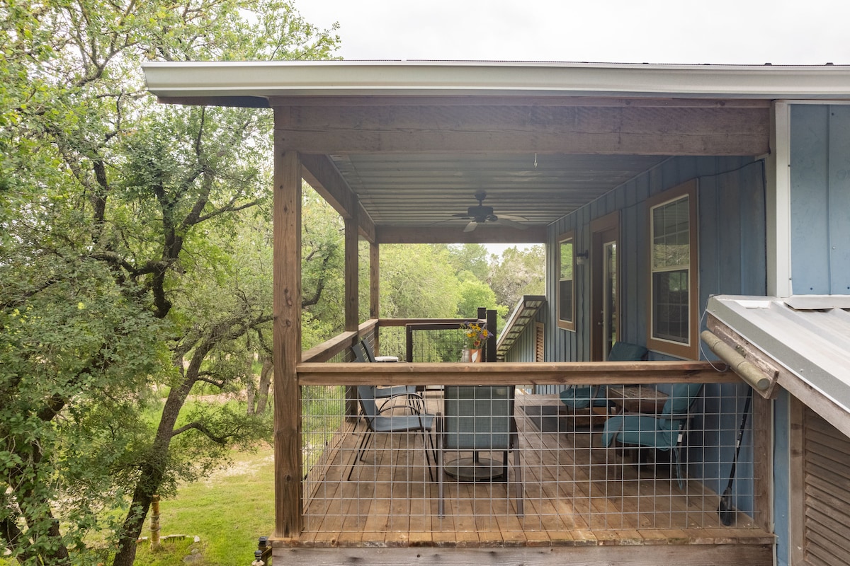 A spacious deck with wooden flooring features seating arrangements, including a table and chairs. Overhead, a ceiling fan provides ventilation while surrounded by lush greenery. Nearby, steps lead down to the ground level, enhancing the outdoor experience.