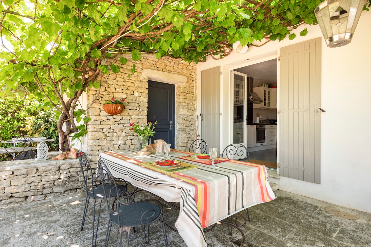 An outdoor dining area is set under a vine-covered trellis, providing shaded seating for six at a rectangular table adorned with a striped tablecloth. Stone walls frame the space, while a door leads into a modern kitchen visible in the background.