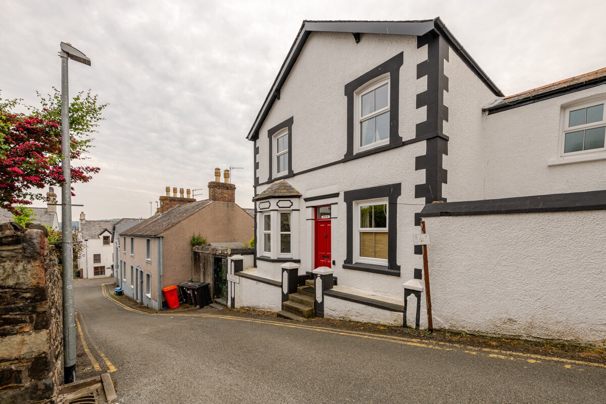The charming exterior of Isallt is displayed, featuring classic white walls with contrasting black trim. A bold red front door adds a pop of color. The house is situated on a quiet street, bordered by greenery and neighboring buildings.