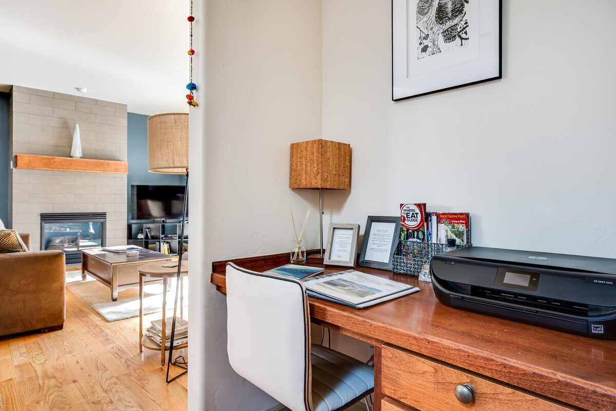 A dedicated workspace is presented, featuring a wooden desk with decorative items and framed documents. A black printer sits neatly beside the desk. Soft lighting from a nearby lamp complements the cozy atmosphere of the adjacent living area with a fireplace.