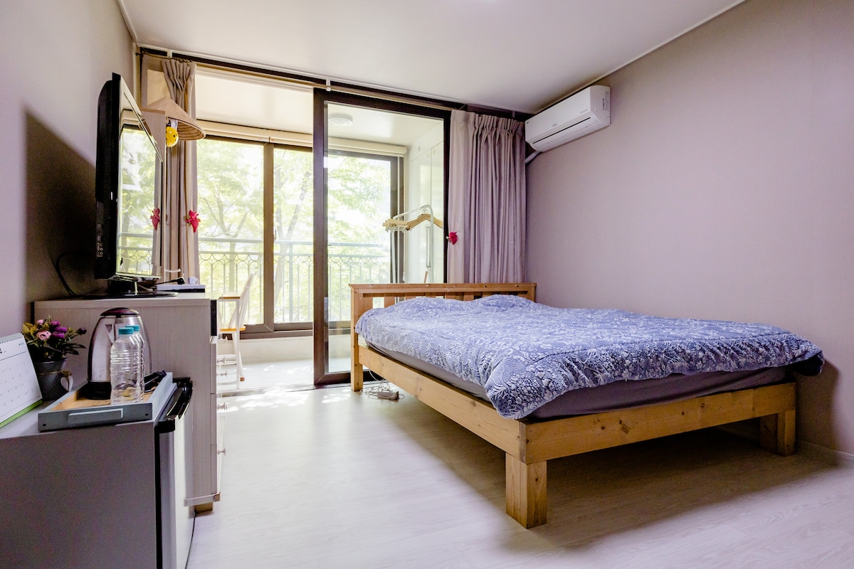 A well-lit guest room features a spacious bed with a patterned blanket, positioned under a window with sheer curtains. A small TV is mounted on the wall, and a desk with a water bottle is visible. Natural light filters in, enhancing the peaceful ambiance.