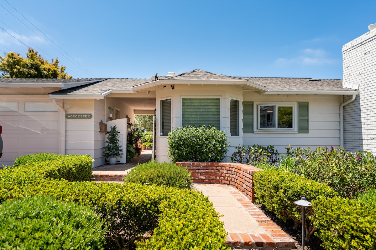 The exterior of a detached master suite is shown, featuring a welcoming entrance surrounded by neatly trimmed hedges and flowering plants. A brick pathway leads towards the covered entryway, which is framed by large windows and decorative shutters under a clear blue sky.