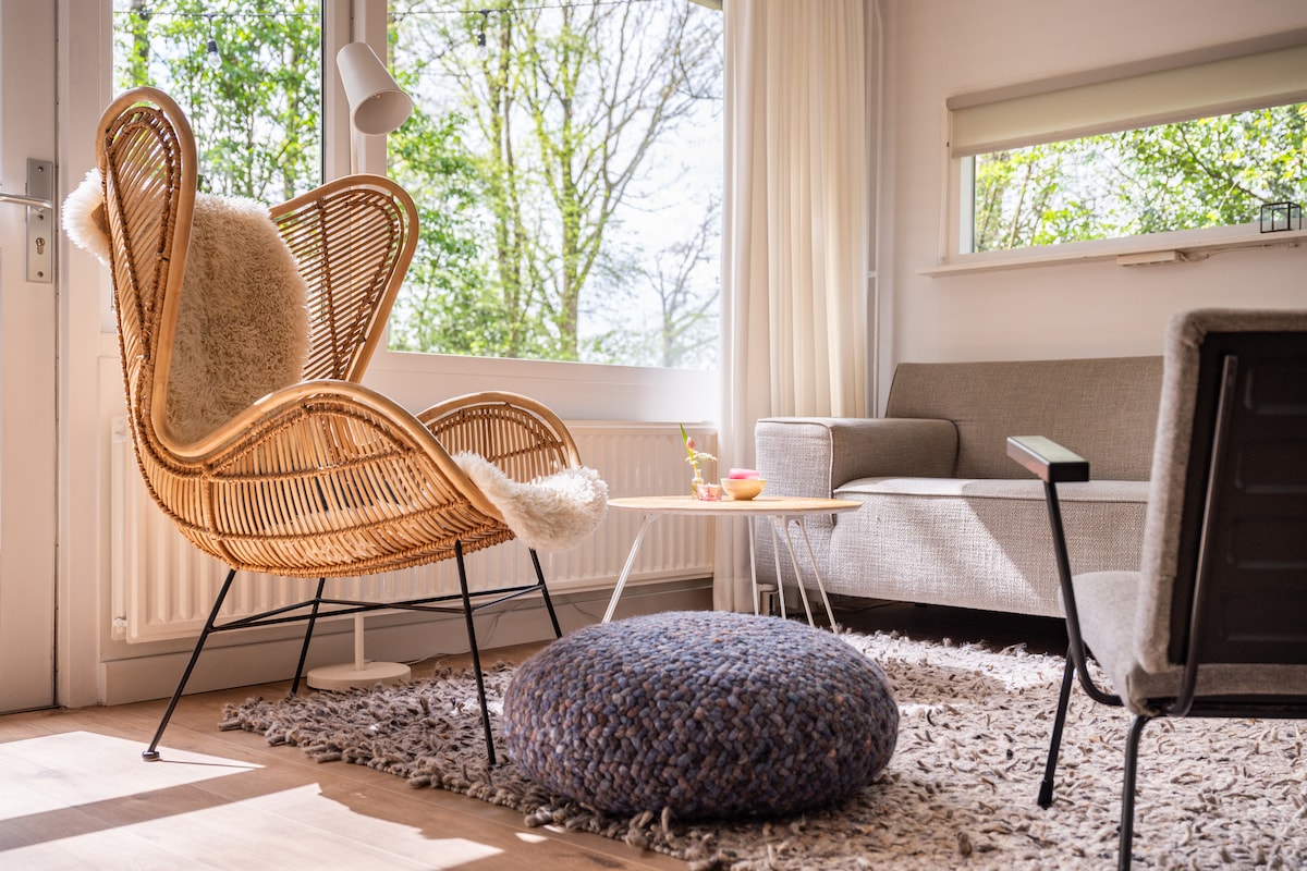 A cozy seating area is highlighted by a rattan chair adorned with a soft throw, a comfortable gray sofa, and a round pouf. Natural light fills the space through large windows, illuminating the light wood flooring and plush rug.