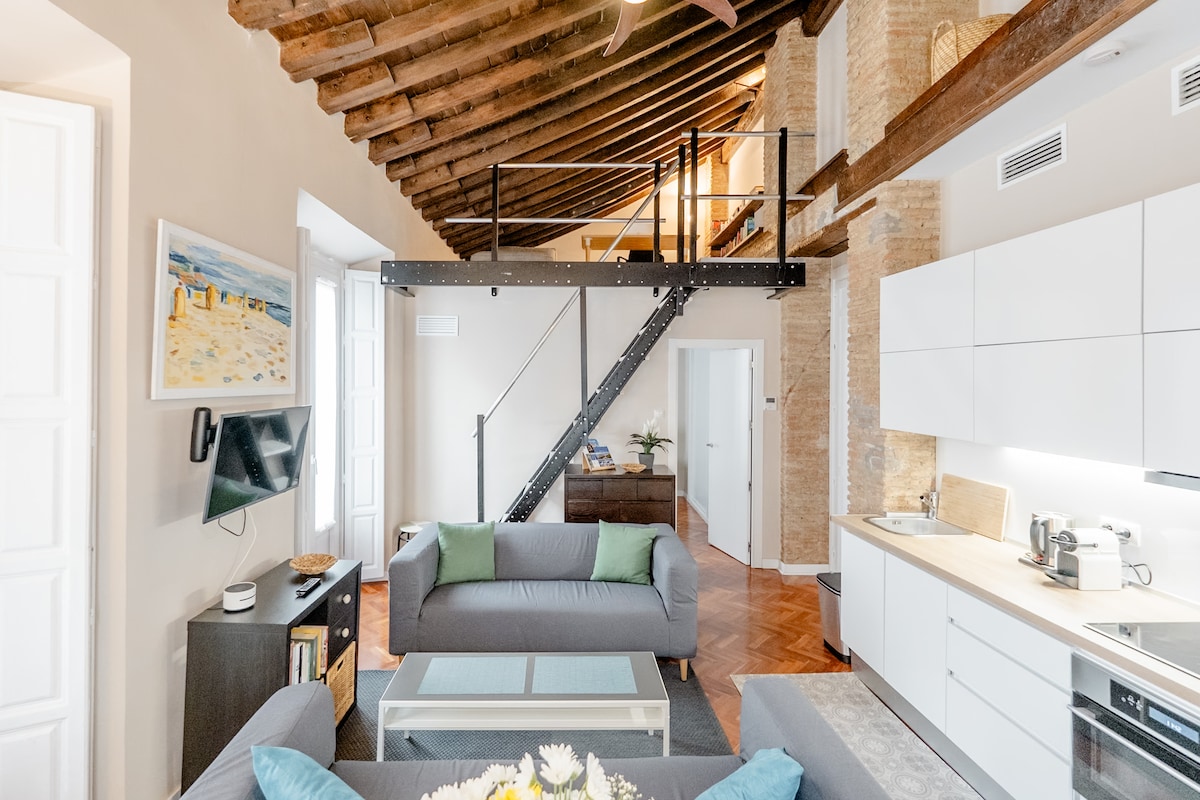 A spacious living area is complemented by high wooden ceilings and exposed brick walls. A staircase leads to a lofted area, while a comfortable seating arrangement is arranged around a central coffee table. Modern kitchen appliances can be seen along one side, providing functional space.