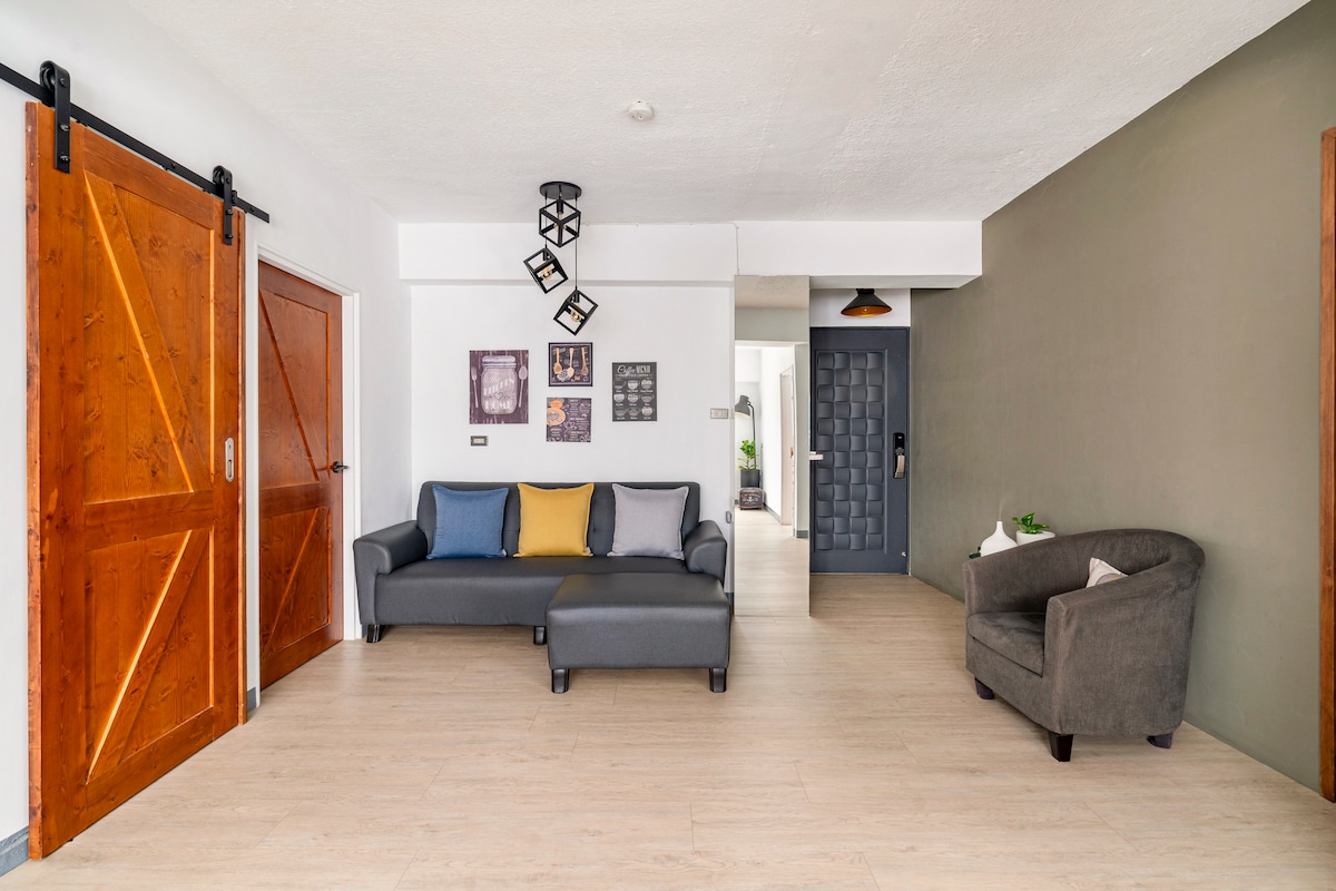 A living area features a comfortable grey sofa with colorful throw pillows, complemented by a matching armchair. Natural light illuminates the room through an open space leading to other areas. Wooden sliding doors offer access to separate rooms, enhancing the layout's openness.