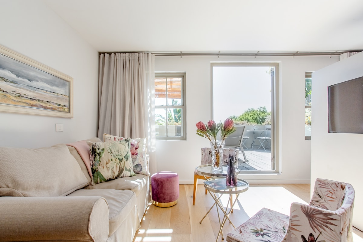 A light-filled living area features a comfortable beige couch adorned with decorative cushions. A small pink ottoman and floral armchairs complement a stylish glass coffee table. Large windows provide views of an outdoor terrace, enhancing the room's openness.