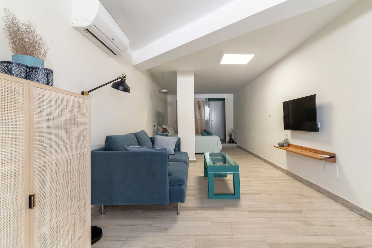 A bright living area is illustrated, featuring a comfortable blue sofa, a glass coffee table, and a wall-mounted television. Soft neutral tones are present on the walls, complemented by natural light entering through a skylight. A separate sleeping area is visible in the background.