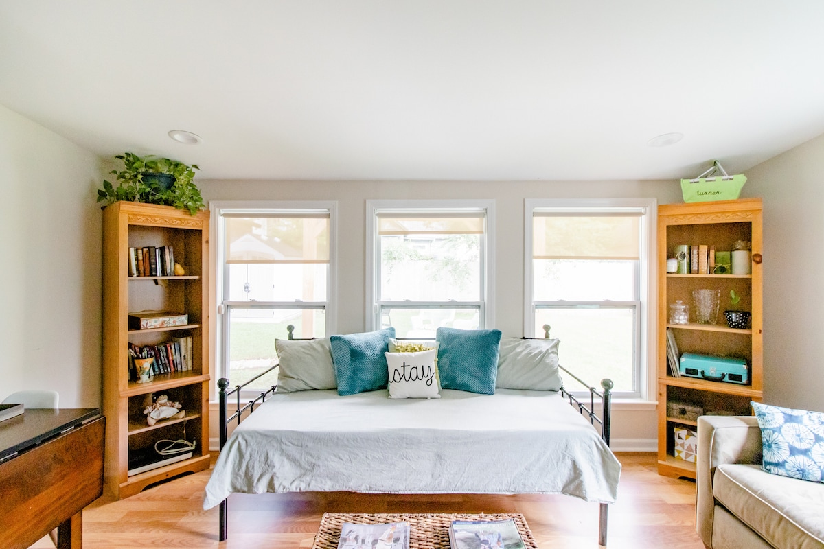 A bright room features a daybed dressed in light linens, complemented by two decorative pillows. Flanking the bed are wooden bookshelves filled with books and decorative items. Natural light enters through three large windows, enhancing the inviting atmosphere of the space.