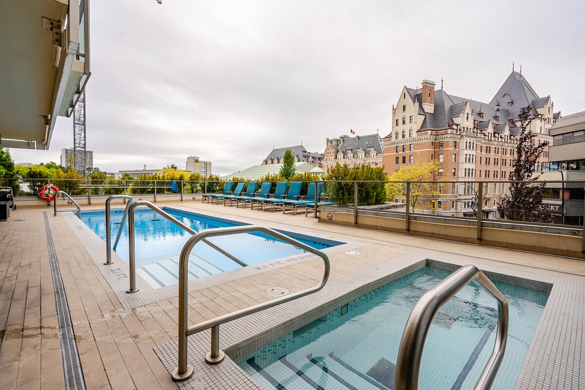 An expansive outdoor pool and hot tub are surrounded by stylish seating loungers. Handrails are installed for safety. The skyline of downtown Victoria serves as a backdrop, featuring buildings with notable architecture. The area is designed for relaxation and leisure.