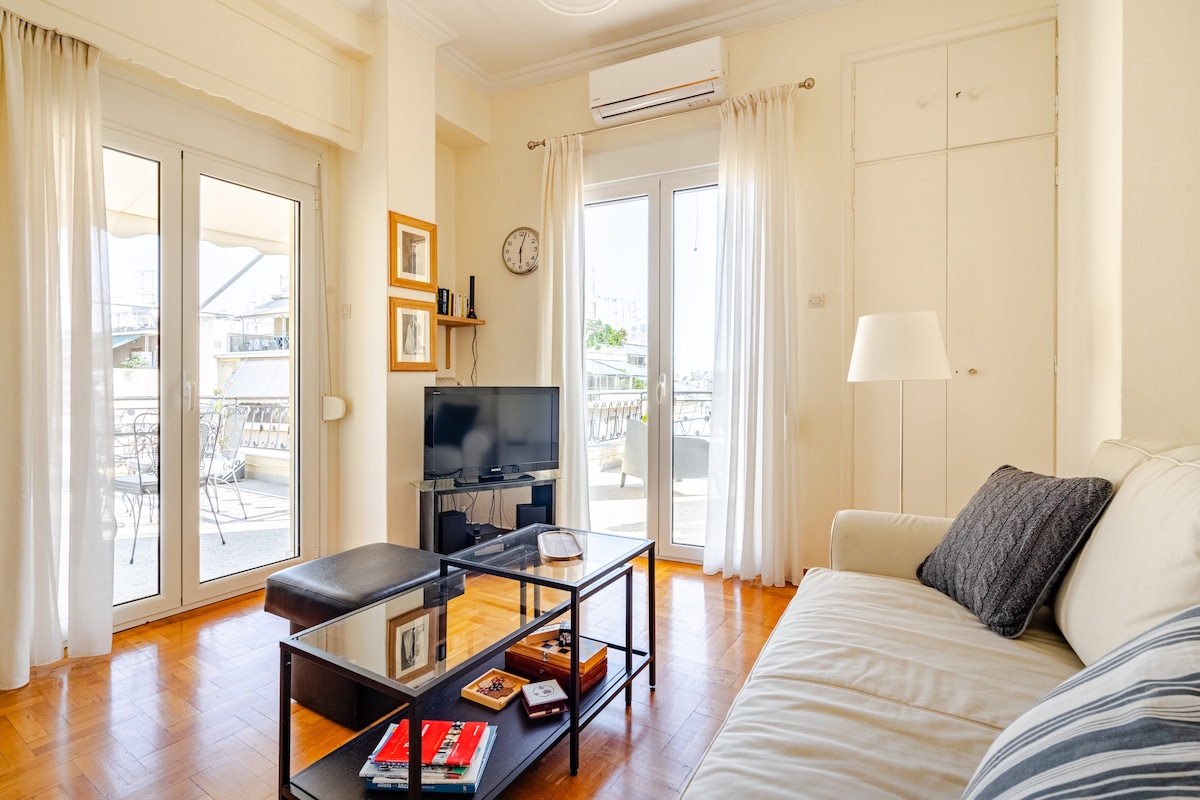 A bright living area features a spacious sofa and a coffee table with various books and magazines. Large glass doors provide access to the balcony, while natural light fills the space. Wall-mounted clock and decorative frames add a welcoming touch.