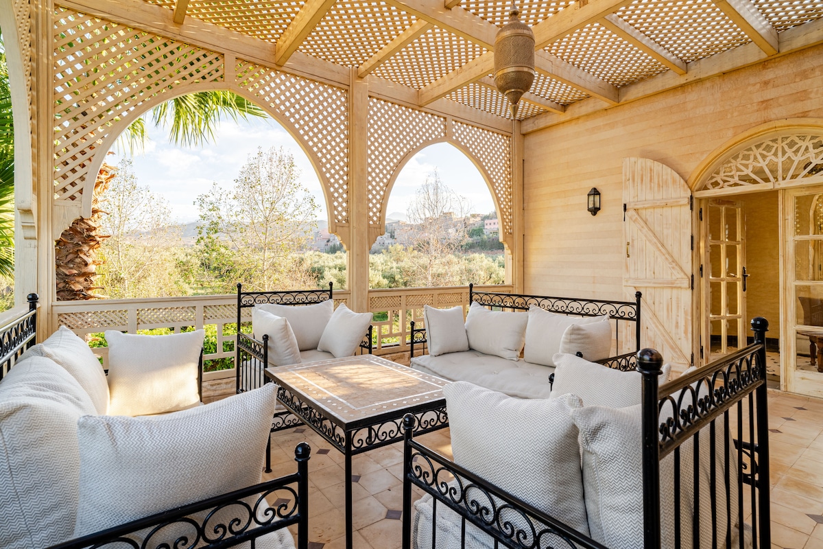 A covered terrace features a series of arched windows allowing natural light to fill the space. Comfortable seating is arranged around a central table, with lush greenery and distant mountains visible through the openings. The traditional design elements are complemented by intricate metalwork.