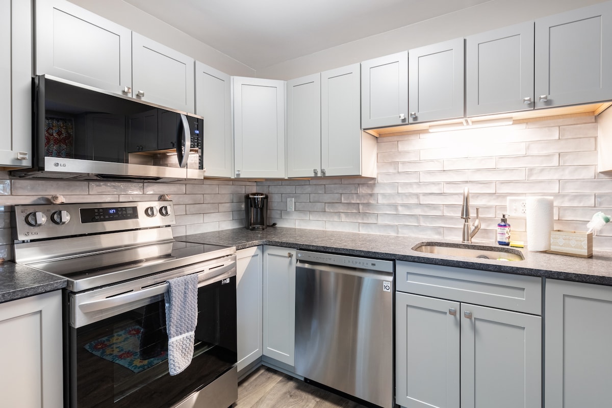 A modern kitchen is equipped with stainless steel appliances, including an oven, microwave, and dishwasher. Light gray cabinets line the walls, contrasting with a textured white tile backsplash. A subtle overhead light enhances the workspace, while a small sink is centrally positioned.