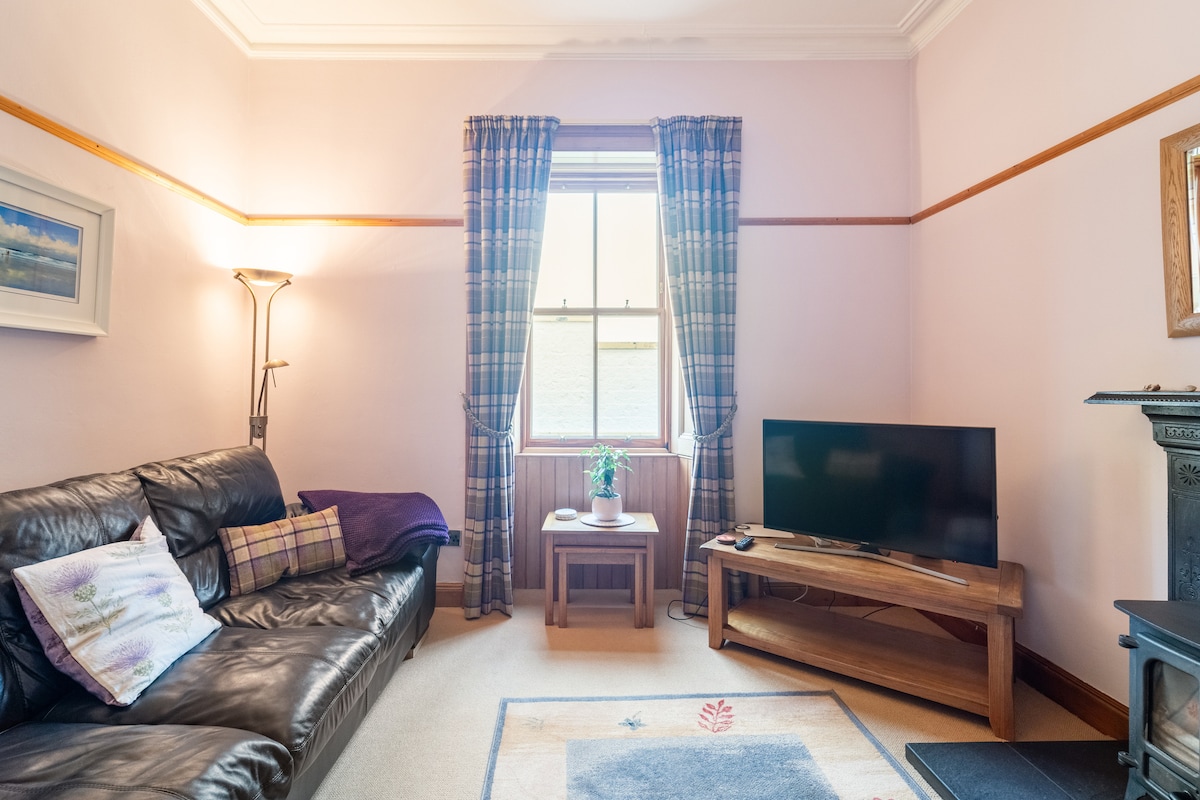 The living room features a leather sofa and a wooden coffee table positioned near a window dressed with plaid curtains. A flat-screen television is placed on a wooden stand, while a decorative plant adds a touch of greenery to the space.