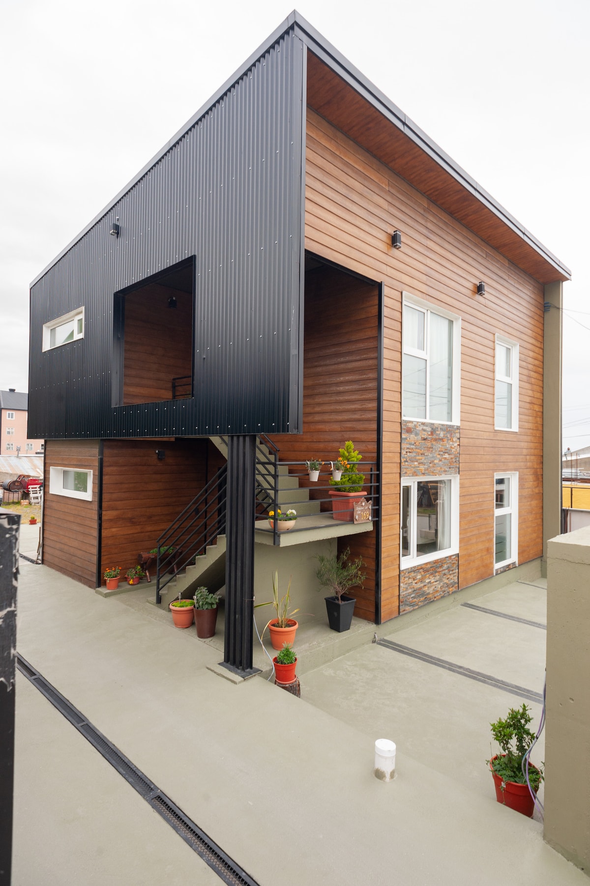 The exterior of the building features a modern design with a combination of wood and metal finishes. A staircase leads to a covered patio area adorned with potted plants. Large windows enhance the structure's contemporary look.