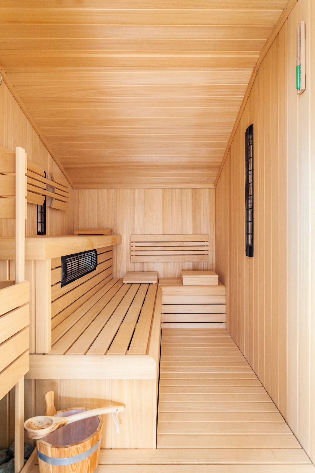 A wooden sauna interior reveals smooth benches along the walls, offering ample seating. The warm-toned wood enhances a natural and calming atmosphere. A bucket and ladle are present, indicating readiness for a traditional sauna experience.