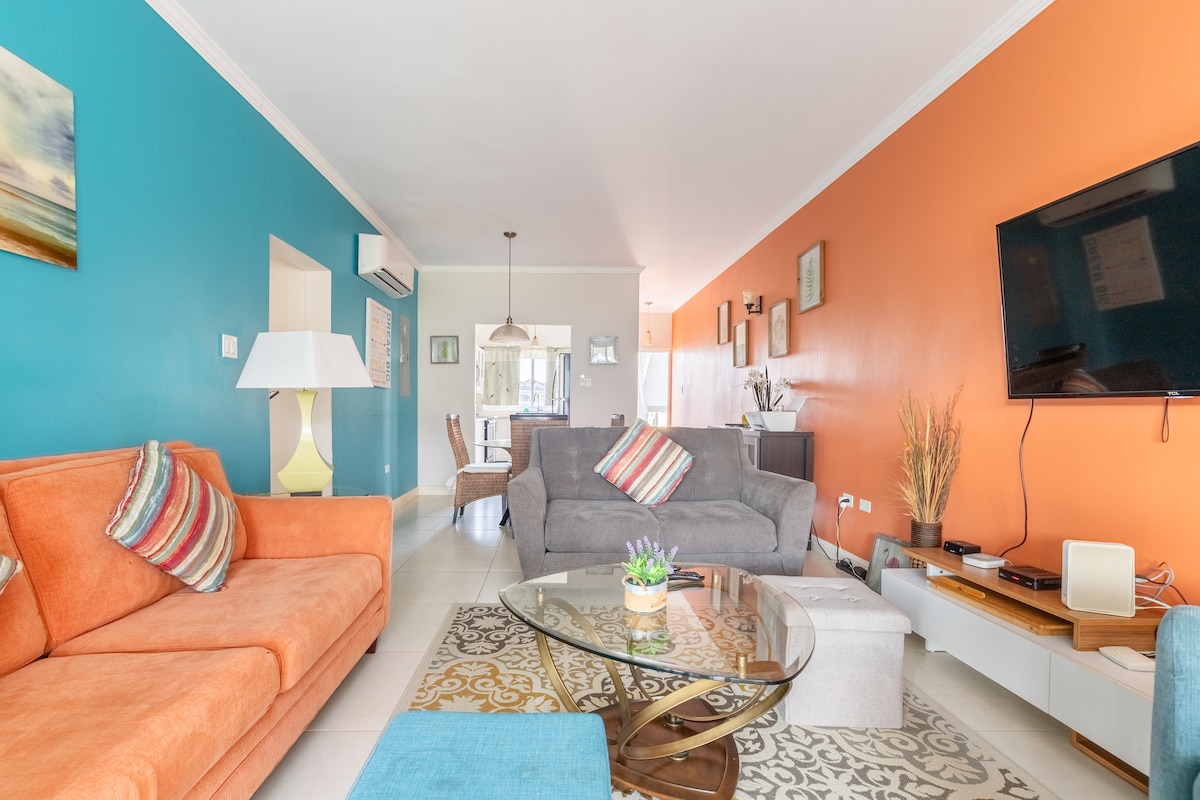 An open living space showcases a blend of vibrant orange and teal walls, with comfortable seating including an orange sofa and a grey couch. A glass coffee table sits at the center, complemented by a decorative arrangement. A large flat-screen TV is mounted on the wall, enhancing the modern aesthetic.