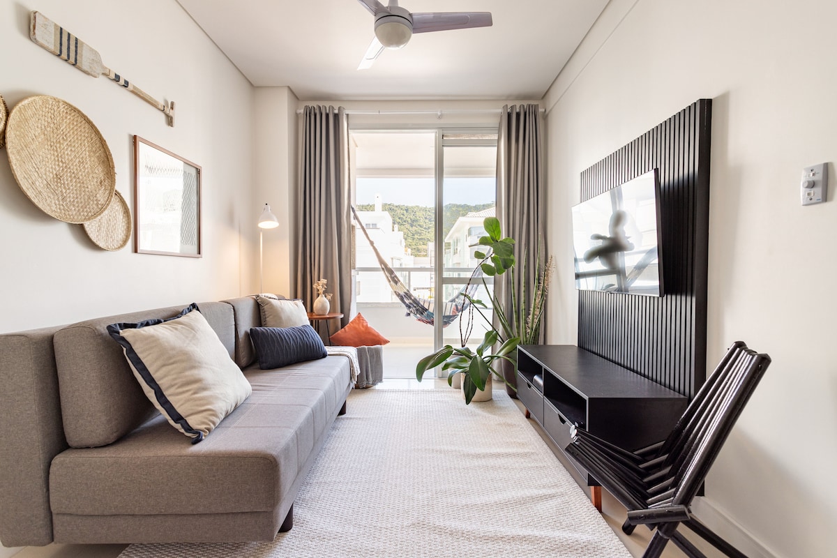 A bright living area features a spacious gray sofa adorned with decorative pillows. A sleek TV unit rests against the wall, accompanied by an indoor plant. Natural light floods the space through large sliding doors leading to a balcony, where a hammock is visible.