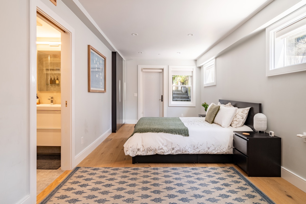 A spacious bedroom features a queen-size bed adorned with a soft green throw. Light enters through multiple windows, including one with a view leading to a staircase. A neutrally toned area rug complements the wooden flooring, while a modern nightstand sits beside the bed.