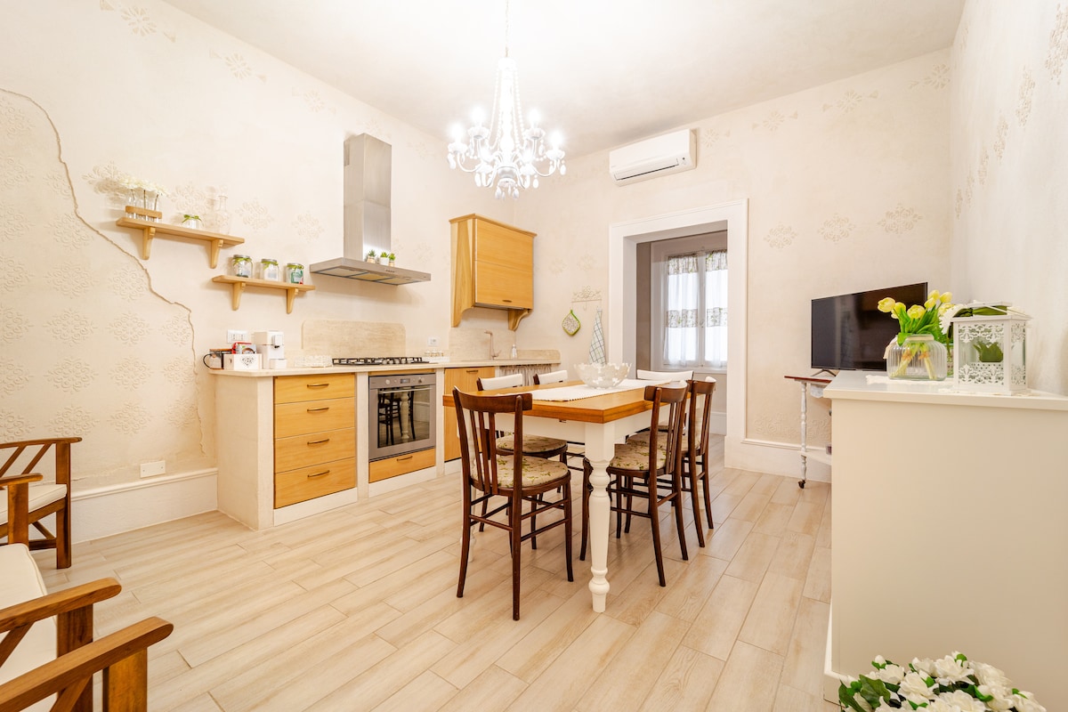 A well-equipped kitchen is highlighted by wooden cabinets and a spacious dining table surrounded by chairs. A modern stove and oven form part of the setup, complemented by a decorative chandelier above. A television and a small shelf are visible in the background.