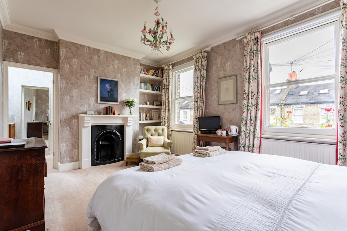 The spacious room contains a kingsize bed adorned with folded towels. A cozy armchair is positioned next to a decorative fireplace. Shelves filled with books line one wall, while two large windows allow natural light to fill the room, framed by floral curtains.