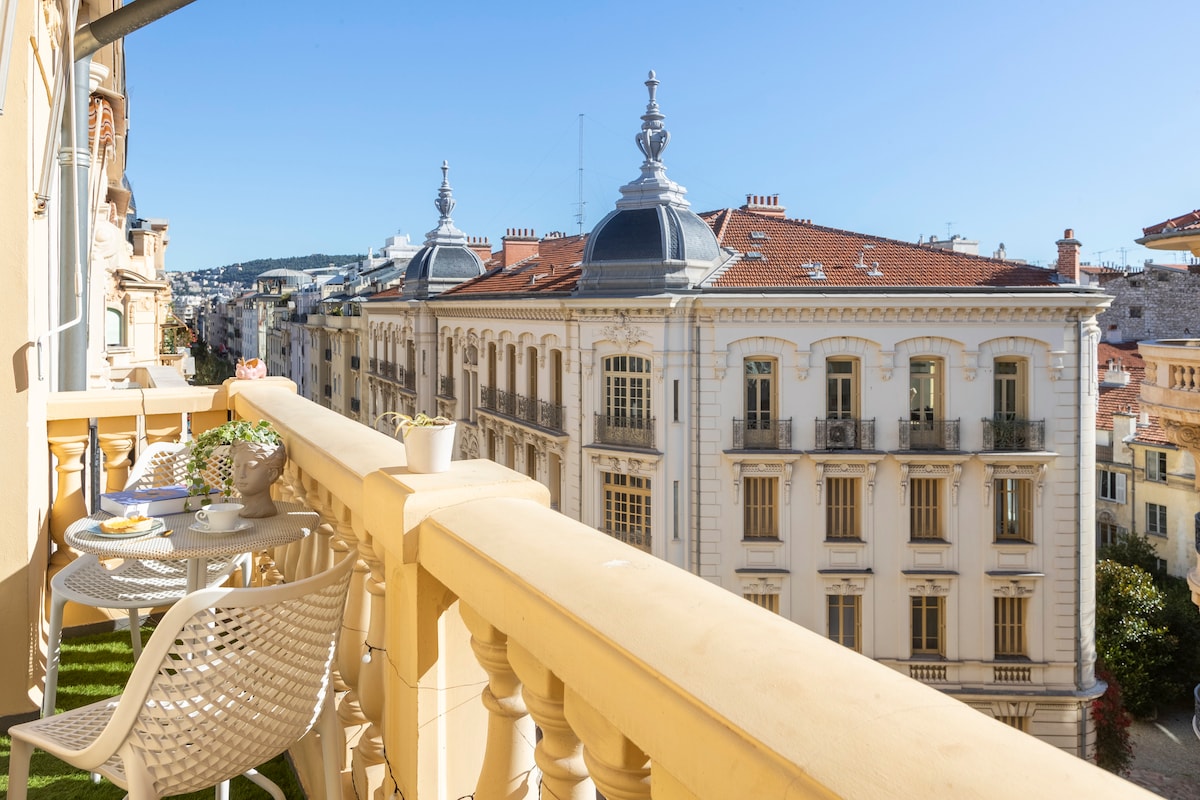 A balcony offers a view of nearby historic buildings, showcasing architectural details and rooftops. Two chairs and a small table are set up on the balcony, accompanied by potted plants. The clear sky enhances the charming atmosphere of the outdoor space.