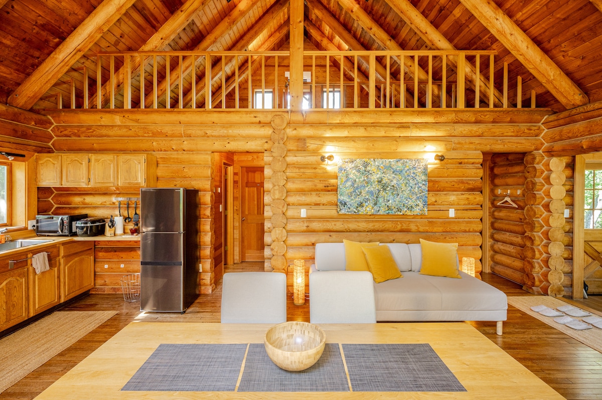 The interior of a log house features high wooden ceilings and warm wood paneling. A spacious dining table is placed at the center, accompanied by a light-colored sofa with yellow cushions. Equipped kitchen appliances and a refrigerator are visible in the background.