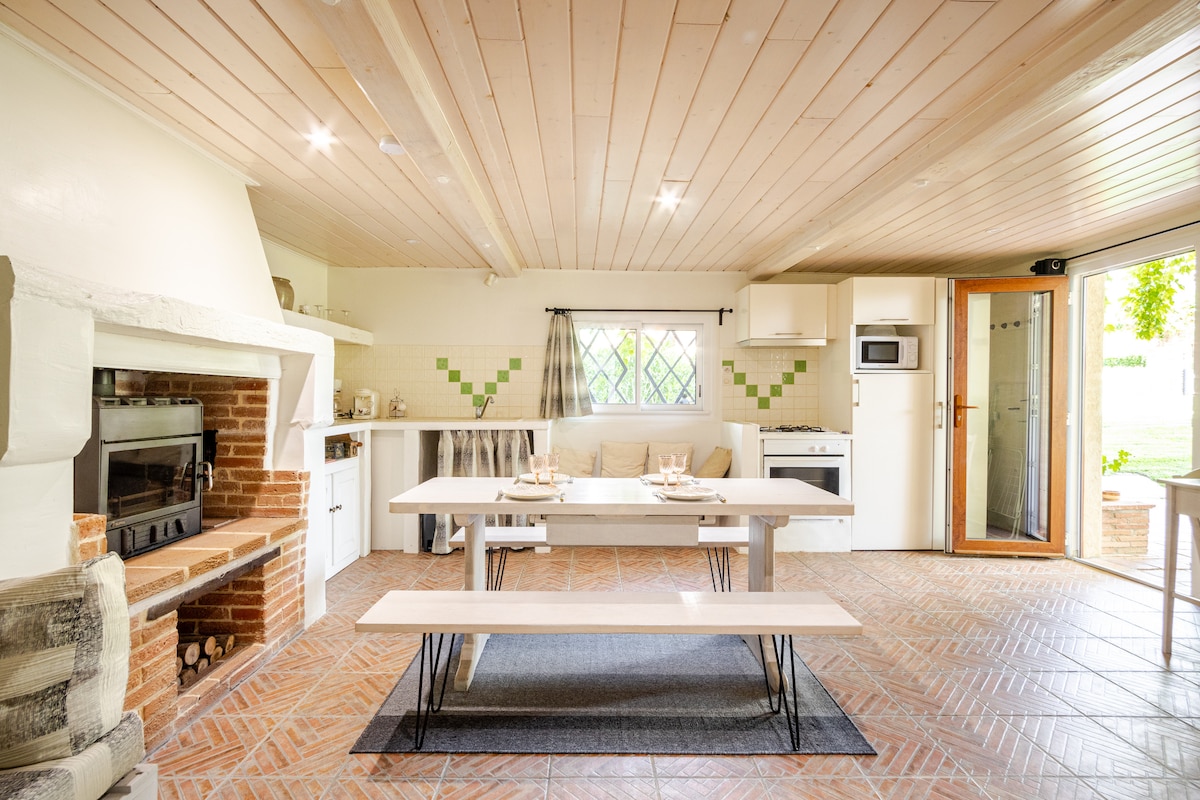 A spacious kitchen and dining area features a rustic design with wooden beams and terracotta tiled flooring. A large dining table is centered in the room, complemented by a well-equipped kitchen that includes an oven, microwave, and ample counter space. Natural light enters through windows overlooking the outdoor space.
