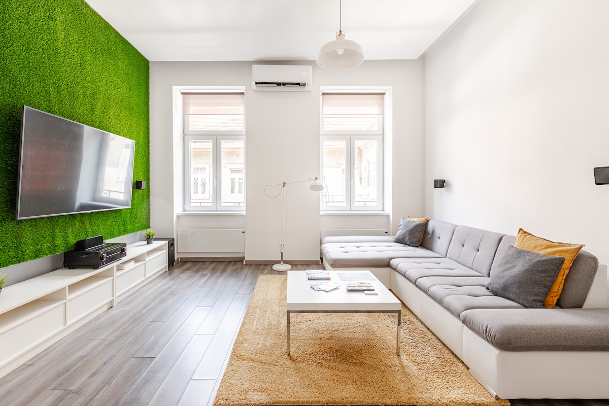 A modern living area features a spacious sectional sofa with gray cushions and orange accents. A large screen TV is mounted on the wall opposite the seating. Two windows allow natural light to fill the room, with a green accent wall adding a touch of freshness.
