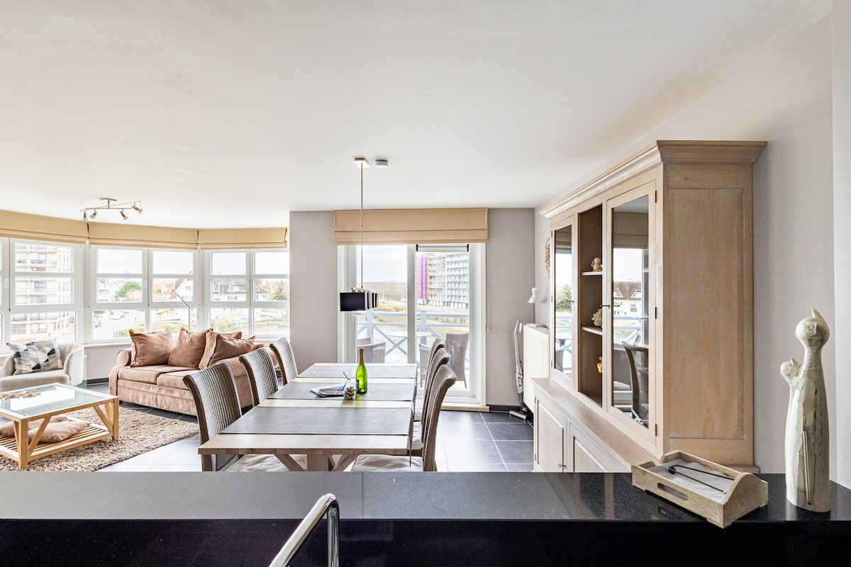 A spacious living area features large windows allowing natural light to fill the space. A comfortable sofa is positioned near a wooden dining table with six chairs. A glass coffee table rests on a soft area rug. The simplicity of the decor provides a relaxed atmosphere.
