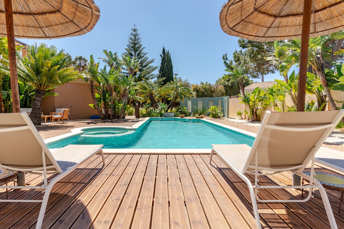 A private pool area features a rectangular pool surrounded by tropical plants and palm trees. Two loungers are positioned on a wooden deck, with a shaded area provided by a thatched umbrella. The inviting blue water reflects sunlight on a clear day.