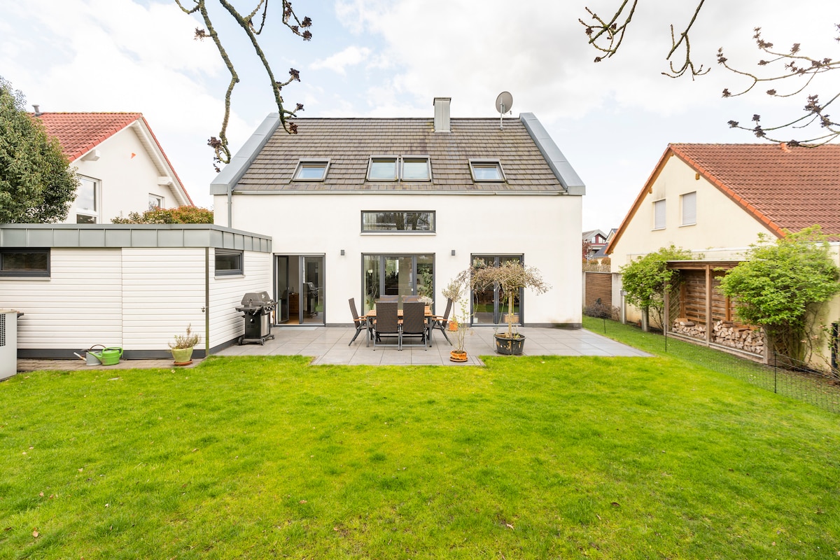 The exterior of a modern home features a spacious patio area with an outdoor dining table and chairs. A gas grill is positioned nearby. The well-maintained lawn is surrounded by greenery, offering a serene environment. Windows are visible on the upper floor, enhancing natural light.