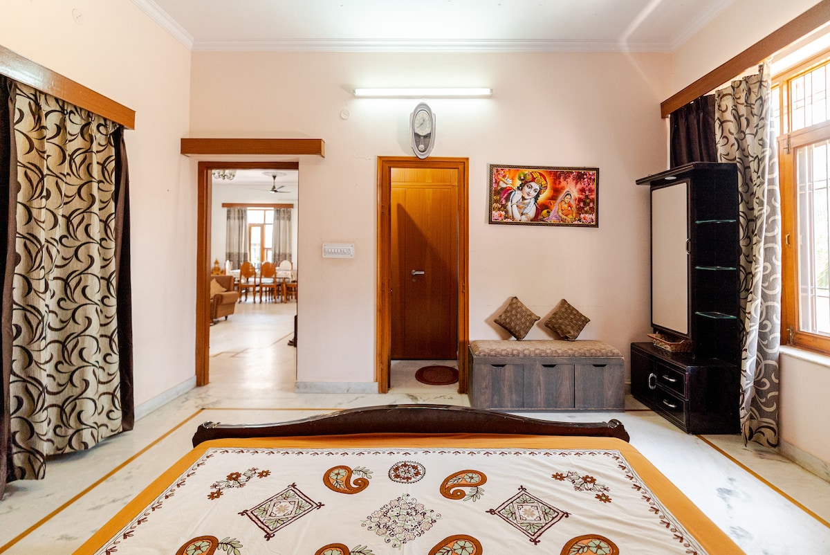The spacious room presents a comfortable bed adorned with an intricate patterned coverlet. A seating area with cushions is visible, along with a dark wood cabinet and wall-mounted television. Sunlight enters through window panes, highlighting the elegant decor and soft color tones around the room.