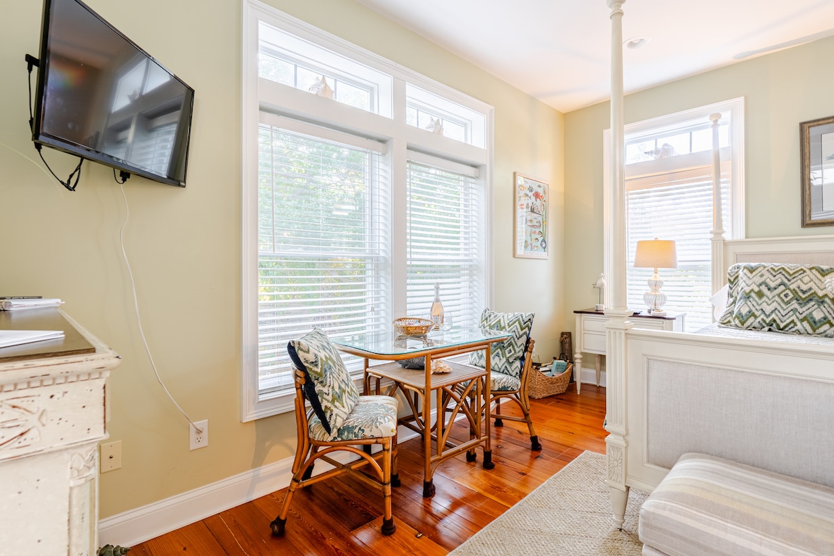 A cozy seating area is visible, featuring a small table and two chairs with patterned cushions. Light filters through double windows, illuminating the warm wooden floor. A flat-screen television is mounted on the wall for entertainment.