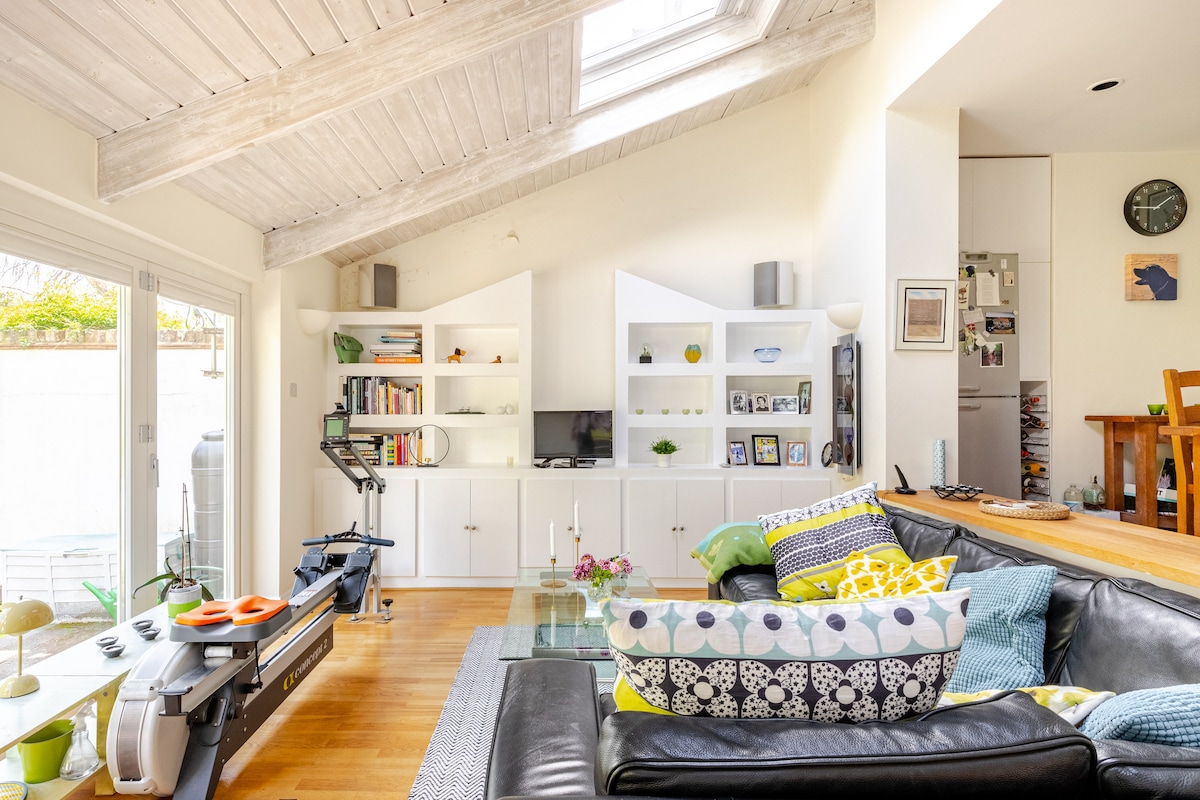 The living area features large skylights that flood the space with natural light. A black sofa with colorful cushions and a glass coffee table adds comfort and style. Shelves are filled with books and decorative items, while a television is positioned for easy viewing.