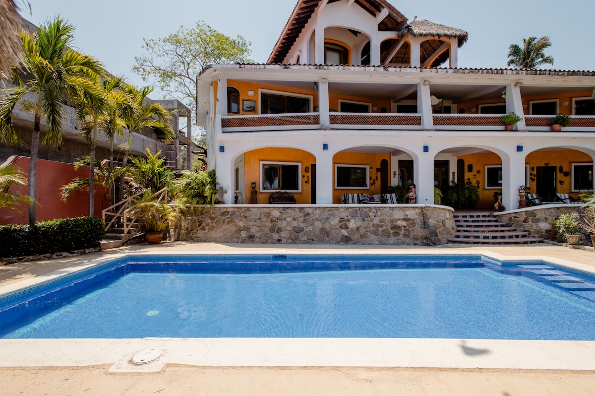 A spacious outdoor area features a clear blue swimming pool surrounded by a stone terrace. The colorful buildings with archways provide a welcoming backdrop, while lush greenery and palm trees enhance the tropical atmosphere. Comfortable seating areas can be seen near the poolside.