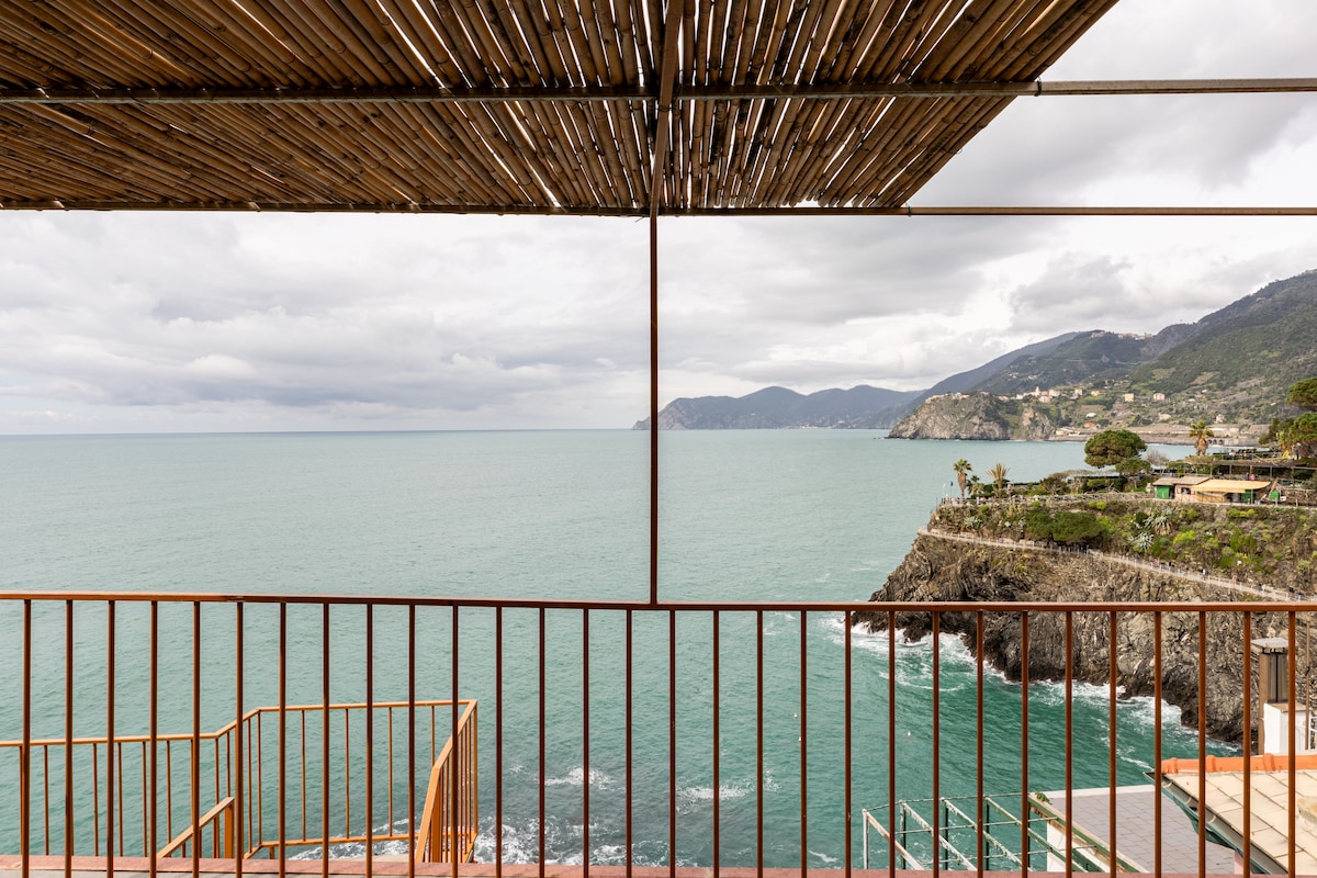 A spacious terrace features a wooden frame providing an unobstructed view of the sea and distant coastline. The tranquil water is complemented by rocky cliffs, with greenery visible on the slopes, creating a serene outdoor space for relaxation.