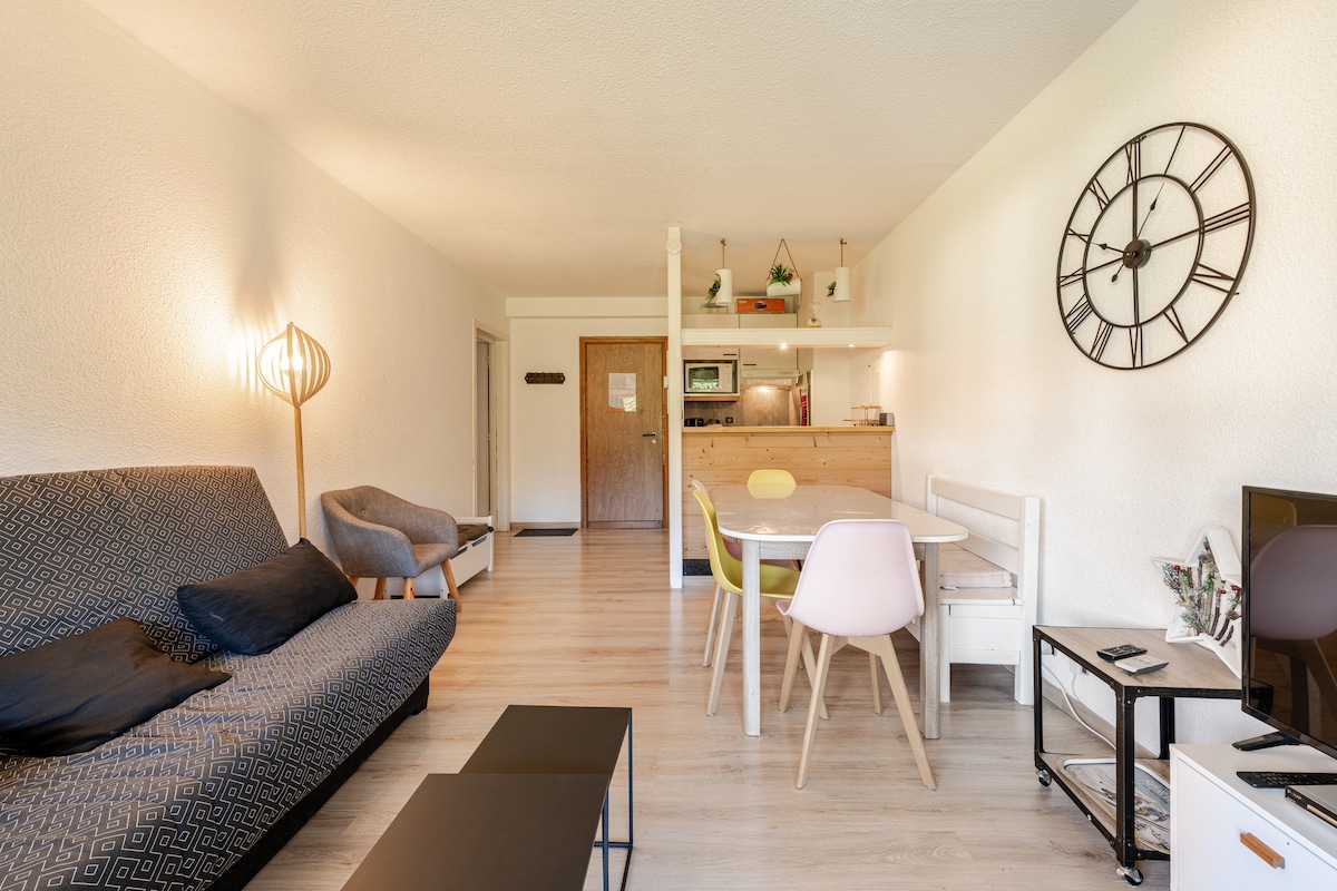 A spacious living and dining area is showcased, featuring a comfortable couch, a dining table with colorful chairs, and a television. The kitchen is visible in the background, with modern appliances and decorative elements. Natural light fills the room, enhancing the welcoming ambiance.