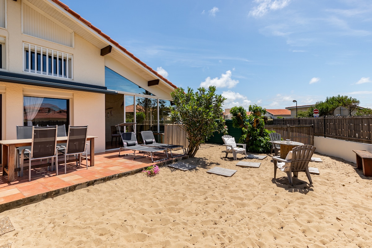 A spacious outdoor area is highlighted, featuring a sand-covered yard. Seating arrangements include a dining table with chairs, and lounge chairs positioned for relaxation. Native plants and a small shrub provide greenery, while a glass sliding door offers access to the indoor space.