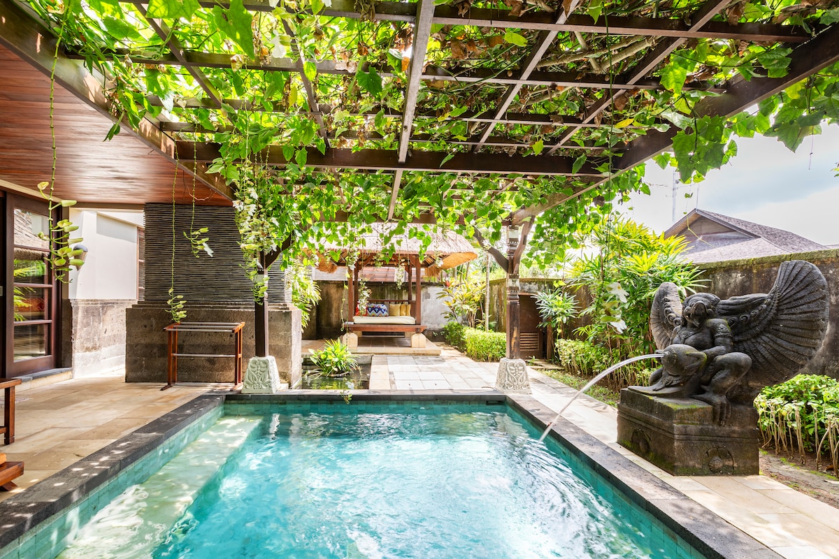 A tranquil private garden features a dip pool surrounded by lush, green vines and a shaded pergola. A traditional gazebo is visible in the background, complemented by a stone sculpture emitting water. Stone pathways and decorative landscaping enhance the peaceful environment.