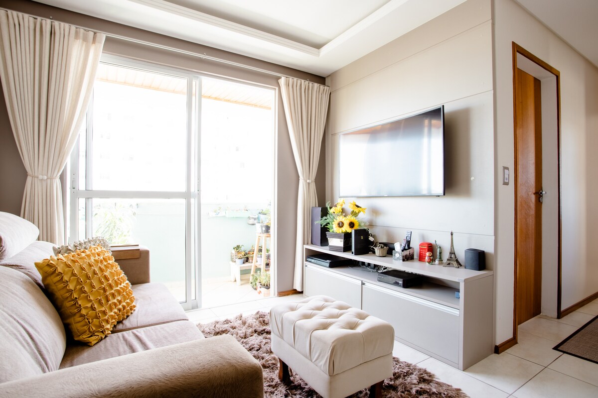 A cozy living area is visible, featuring a light-colored sofa adorned with decorative cushions. A flat-screen TV is mounted on the wall above a modern media console, which holds various decorative items. Natural light streams in through the sliding glass door, illuminating the space.