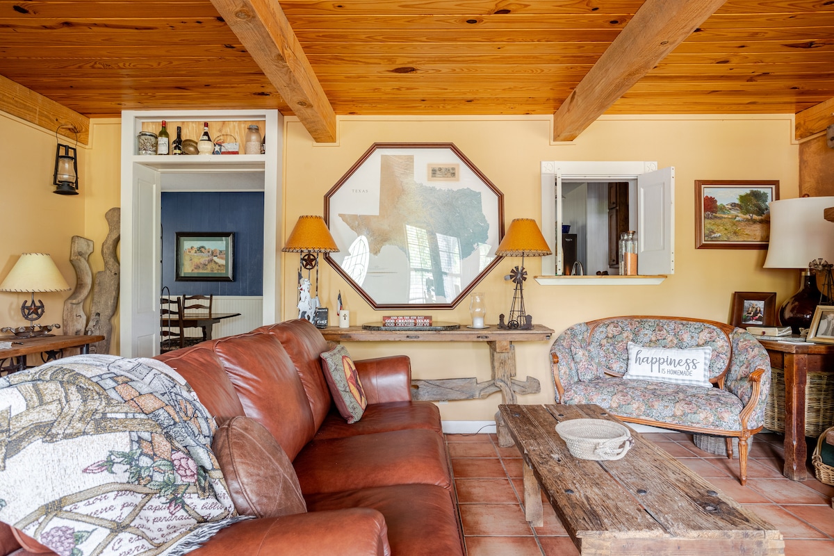 A cozy living area features a brown leather sofa with decorative pillows and a floral armchair. A wooden coffee table sits in the center, surrounded by warm wood beams and light-colored walls. Natural light from windows illuminates the space, highlighting various personal decor items.