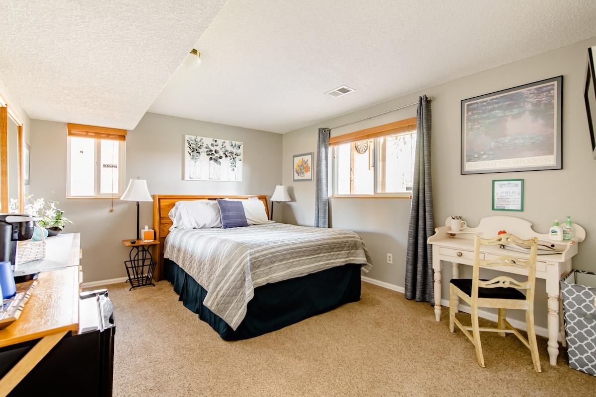 A spacious bedroom features a queen temperpedic bed with neutral bedding and an accent throw. Natural light filters in through two windows, framed by light curtains. A small desk and chair are positioned beside a charging station, while a Keurig coffee maker is located on a nearby side table.