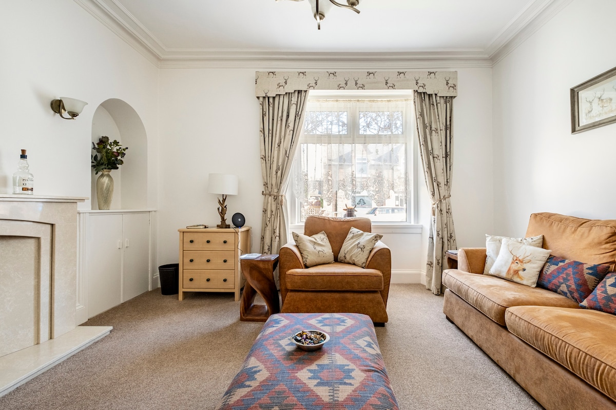 A comfortable living area features a tan sofa and a matching armchair, complemented by decorative cushions. A coffee table sits at the center, adorned with a small bowl. Natural light filters through a large window, enhancing the cozy atmosphere of the space.