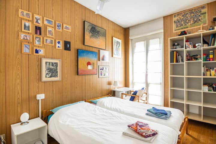 Поглед кон спалната соба, со кревети, работна маса, прозорец во паркот, тавански вентилатор.